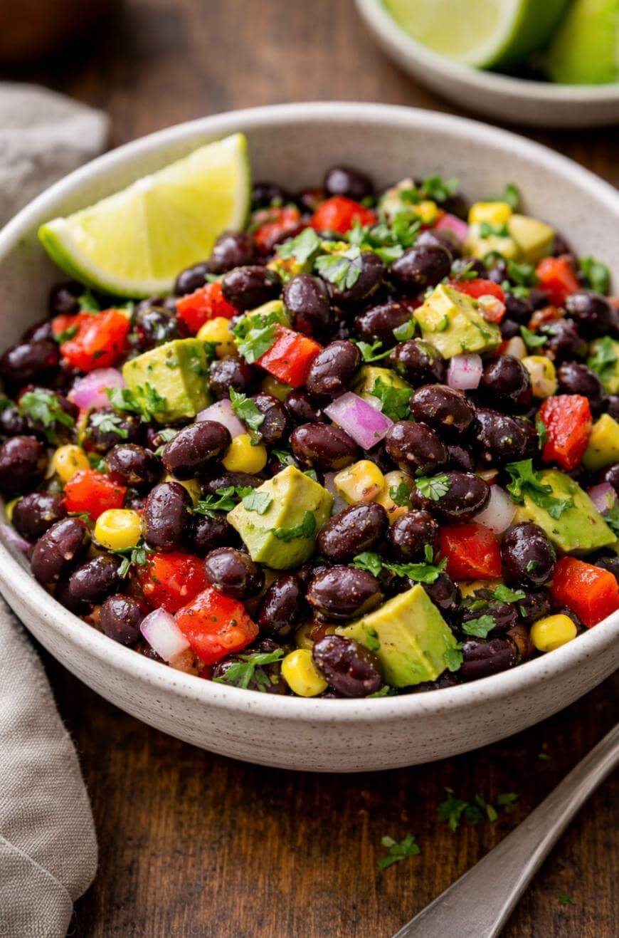 A bowl of black bean salad with diced avocado, red bell pepper, corn, red onion, and chopped cilantro, garnished with a lime wedge, on a wooden table.