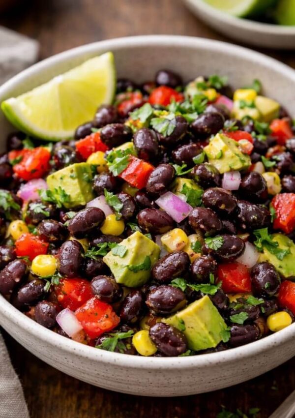 A bowl of black bean salad with black beans, diced avocado, corn, chopped tomatoes, red onion, cilantro, and a lime wedge on the side.