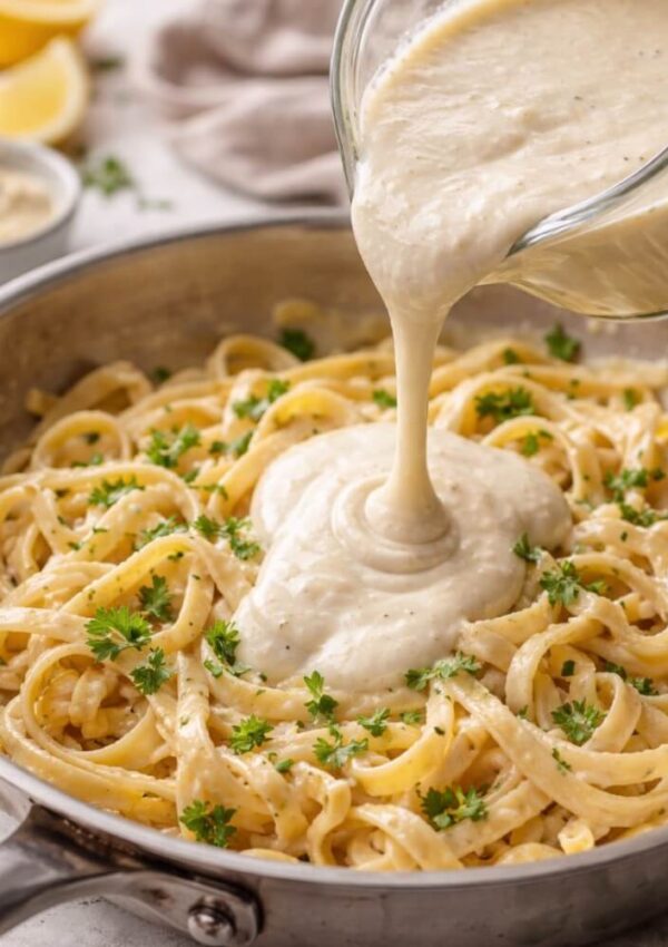 A creamy white sauce is being poured from a glass container onto a pan of fettuccine pasta, garnished with chopped parsley. Lemons and a bowl of sauce are visible in the background.