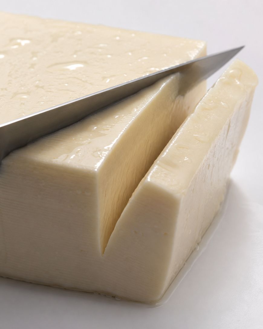 A close-up of a knife slicing through a large, rectangular block of butter on a white surface. The butter appears smooth and creamy, with a glossy texture.