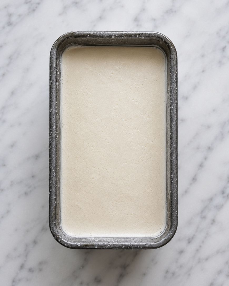 A rectangular metal pan filled with smooth, creamy white ice cream sits on a white marble surface, viewed from above.