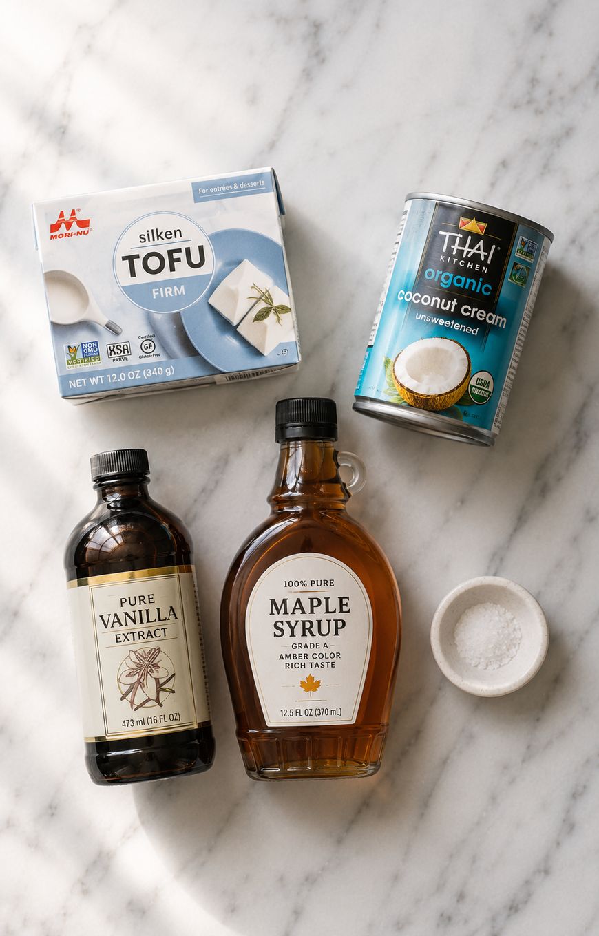 A flat lay of silken tofu, a can of coconut cream, bottles of vanilla extract and maple syrup, and a small bowl of salt on a light surface.