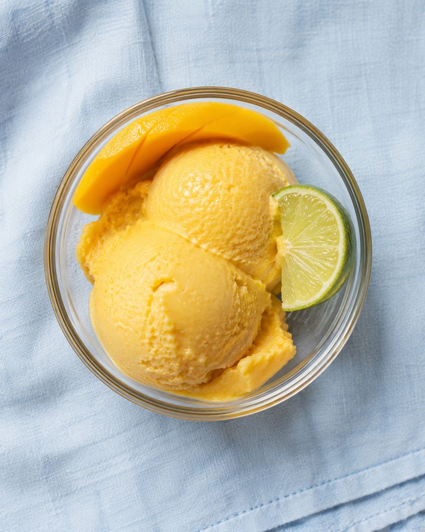A glass bowl with two scoops of mango sorbet, garnished with a mango slice and a lime wedge, placed on a light blue fabric background.