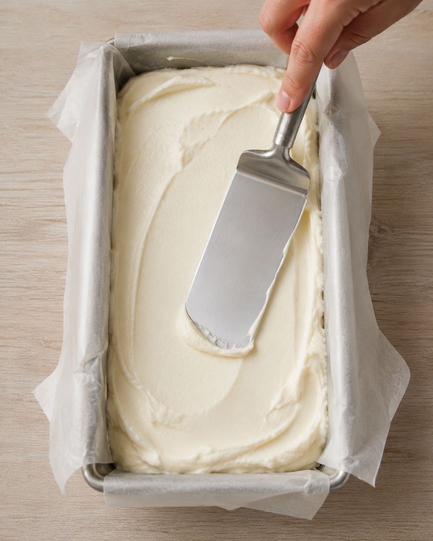 A hand uses a metal spatula to smooth creamy white batter in a parchment-lined rectangular baking pan on a light wooden surface.