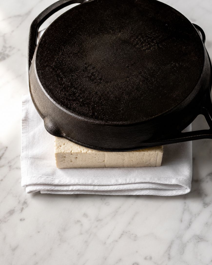 A cast iron skillet presses a block of tofu wrapped in a white cloth on a marble surface.
