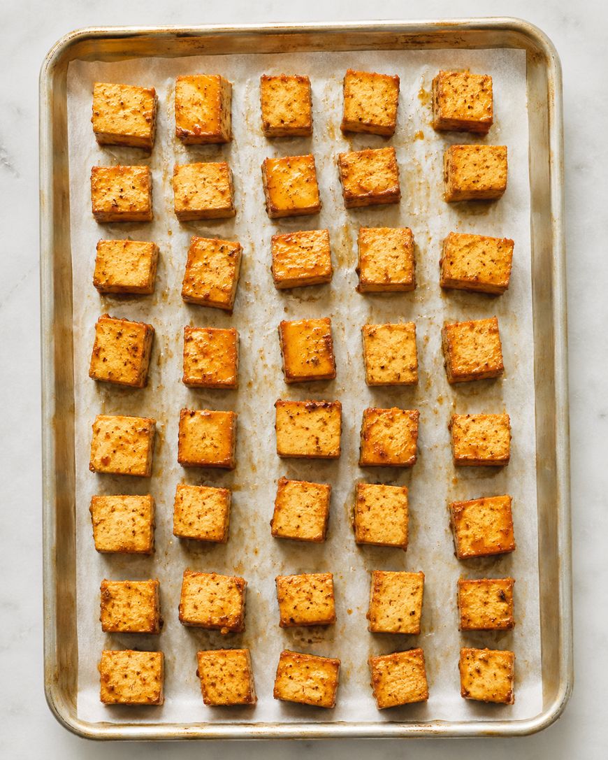 A baking sheet with rows of evenly spaced, golden brown, seasoned tofu cubes on parchment paper, seen from above on a light surface.