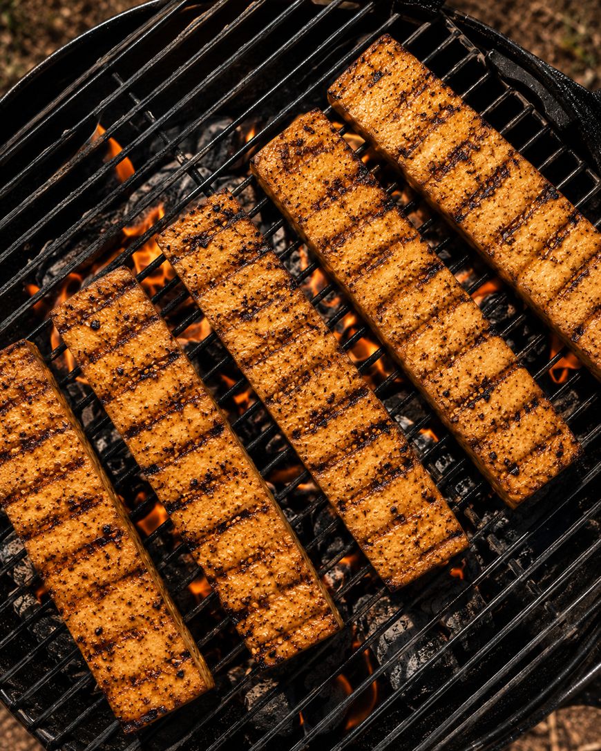 Six seasoned tofu slabs with grill marks are cooking over hot charcoal and flames on a barbecue grill.
