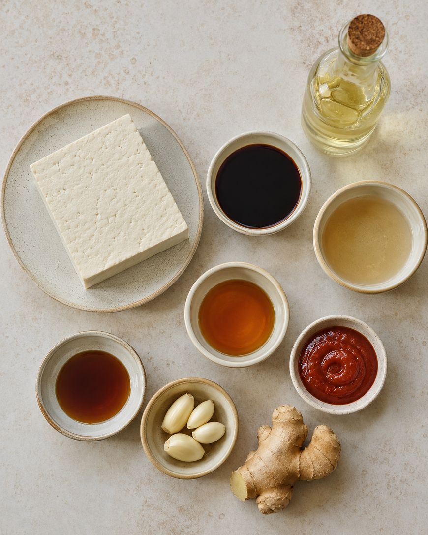 A block of tofu on a plate, surrounded by small bowls of soy sauce, rice vinegar, sesame oil, hot sauce, maple syrup, garlic cloves, ginger root, and a bottle of oil on a neutral surface.