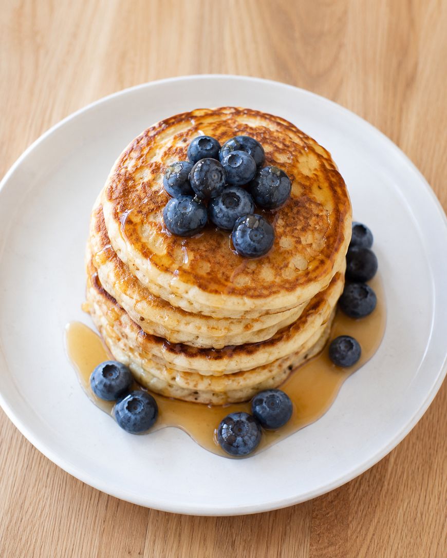 A stack of three pancakes topped with fresh blueberries and drizzled with syrup, served on a white plate with extra blueberries and syrup around the base.