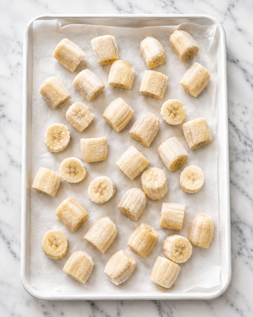 A baking sheet lined with parchment paper holds evenly spaced, sliced pieces of banana, ready for freezing on a marble countertop.