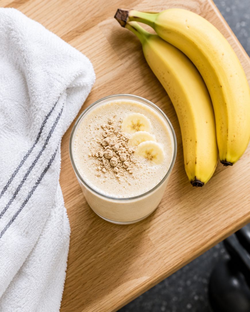 A glass of banana smoothie topped with banana slices and powder sits on a wooden surface next to two whole bananas and a white towel with a dark stripe.