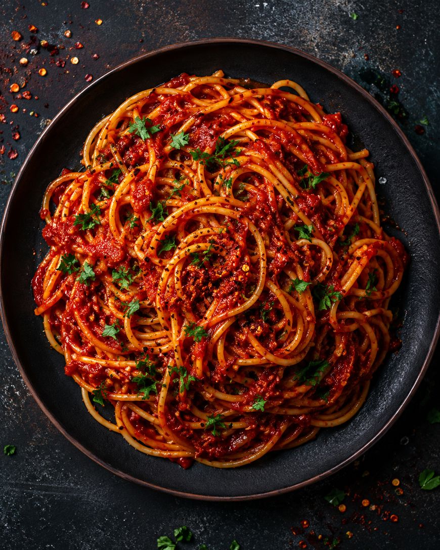 A dark plate filled with spaghetti topped with rich red tomato sauce, garnished with chopped parsley and red pepper flakes, sits on a dark textured surface.