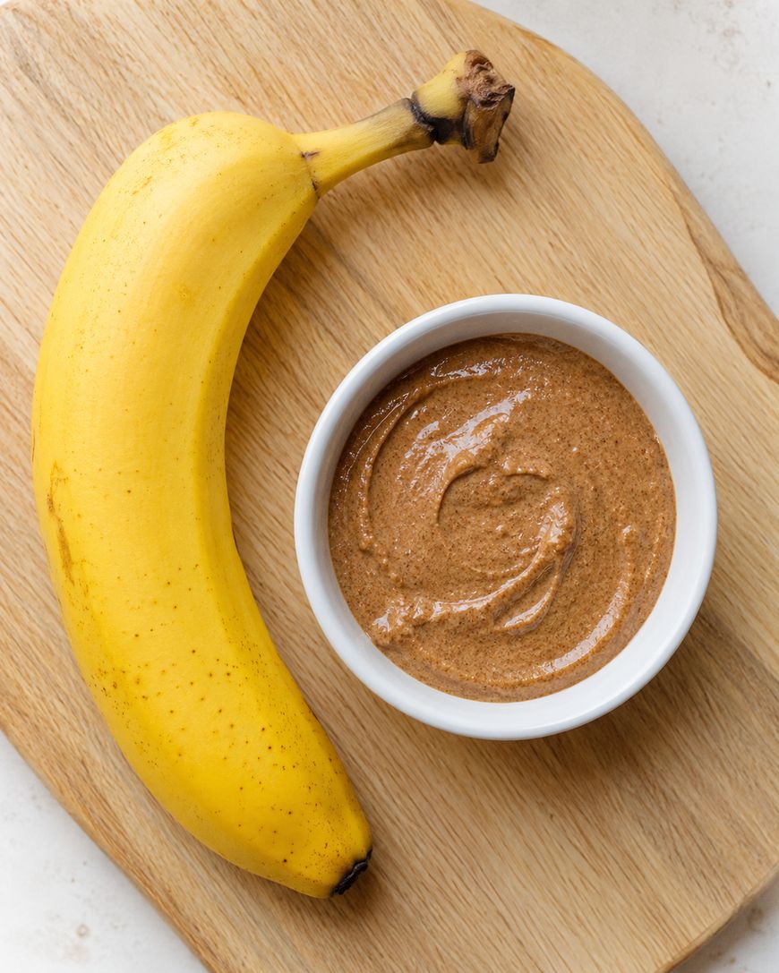 A ripe banana and a small white bowl of almond butter are placed on a wooden cutting board.