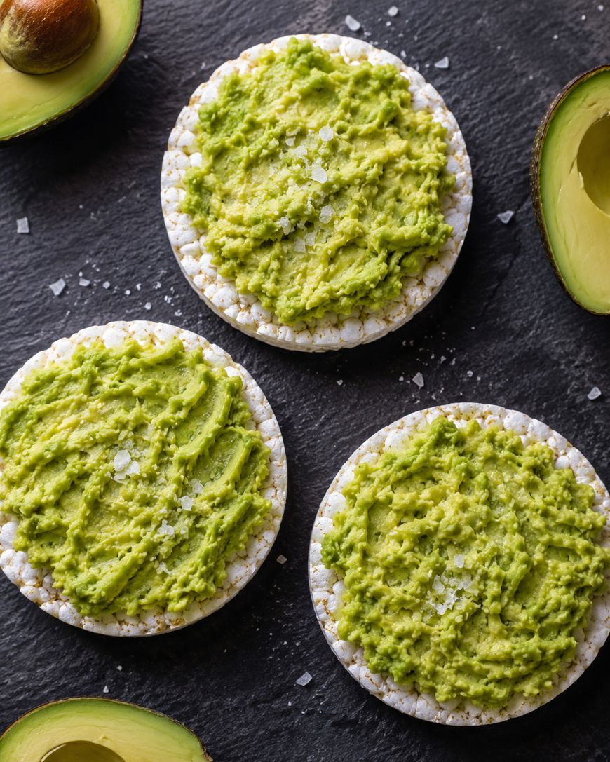Three rice cakes topped with mashed avocado and sprinkled with sea salt are arranged on a dark surface, surrounded by halved and whole avocados.