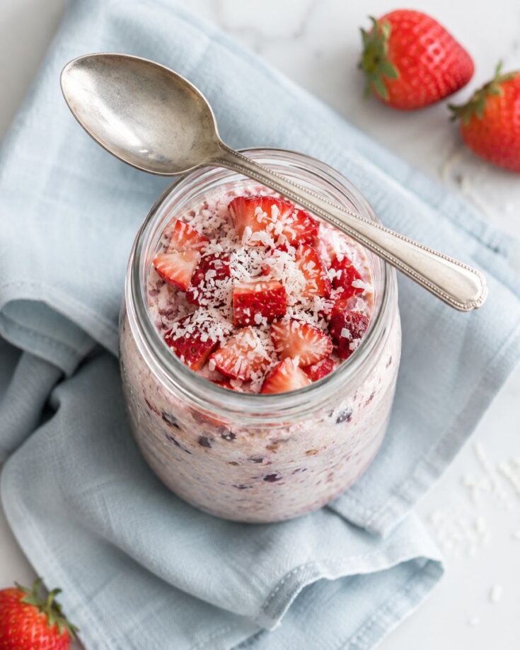 A jar of overnight oats topped with chopped strawberries and shredded coconut sits on a folded blue cloth napkin, with a spoon resting on top and fresh strawberries nearby.