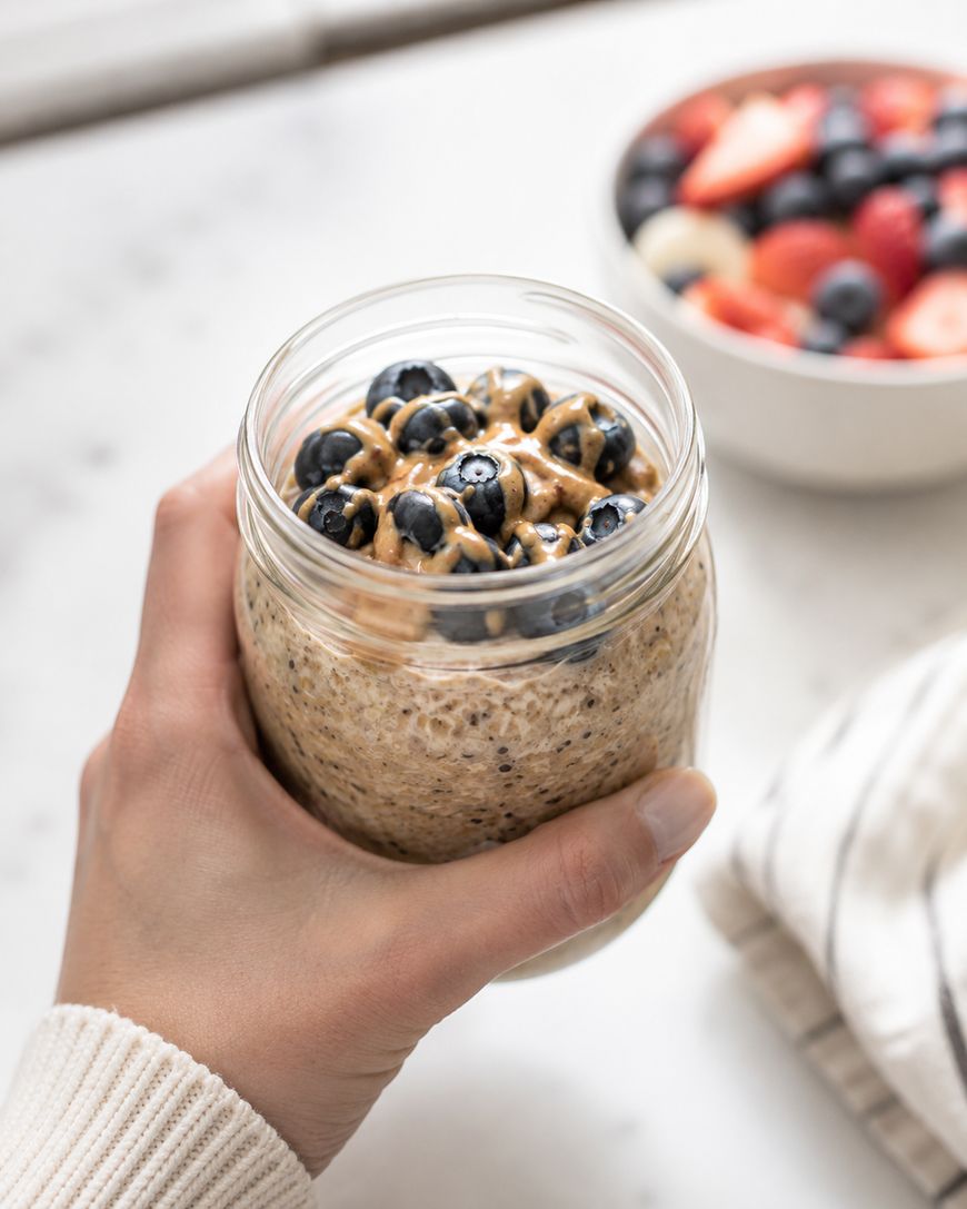 A hand holds a jar of overnight oats topped with fresh blueberries and a drizzle of nut butter. In the background, there is a bowl of mixed berries and a folded cloth.