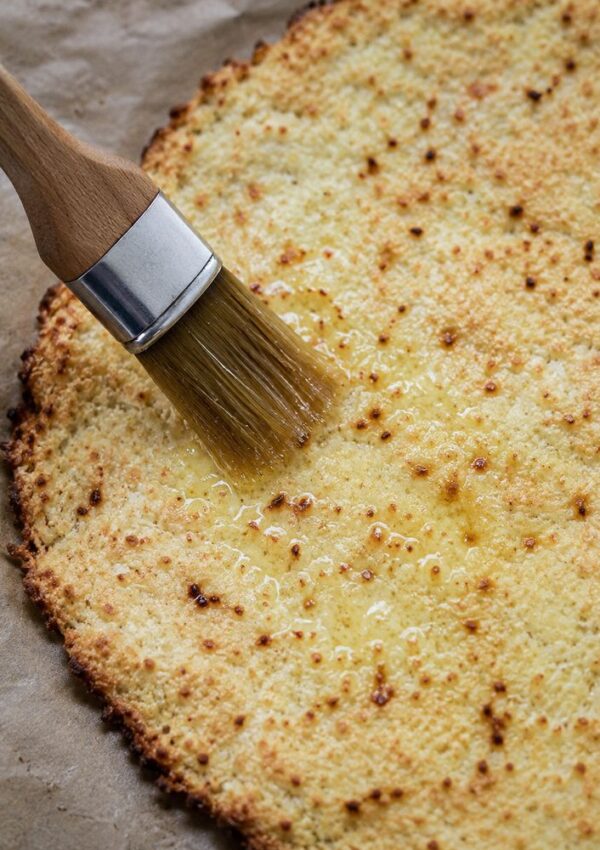 A pastry brush spreads melted butter over a golden, baked flatbread crust on parchment paper.