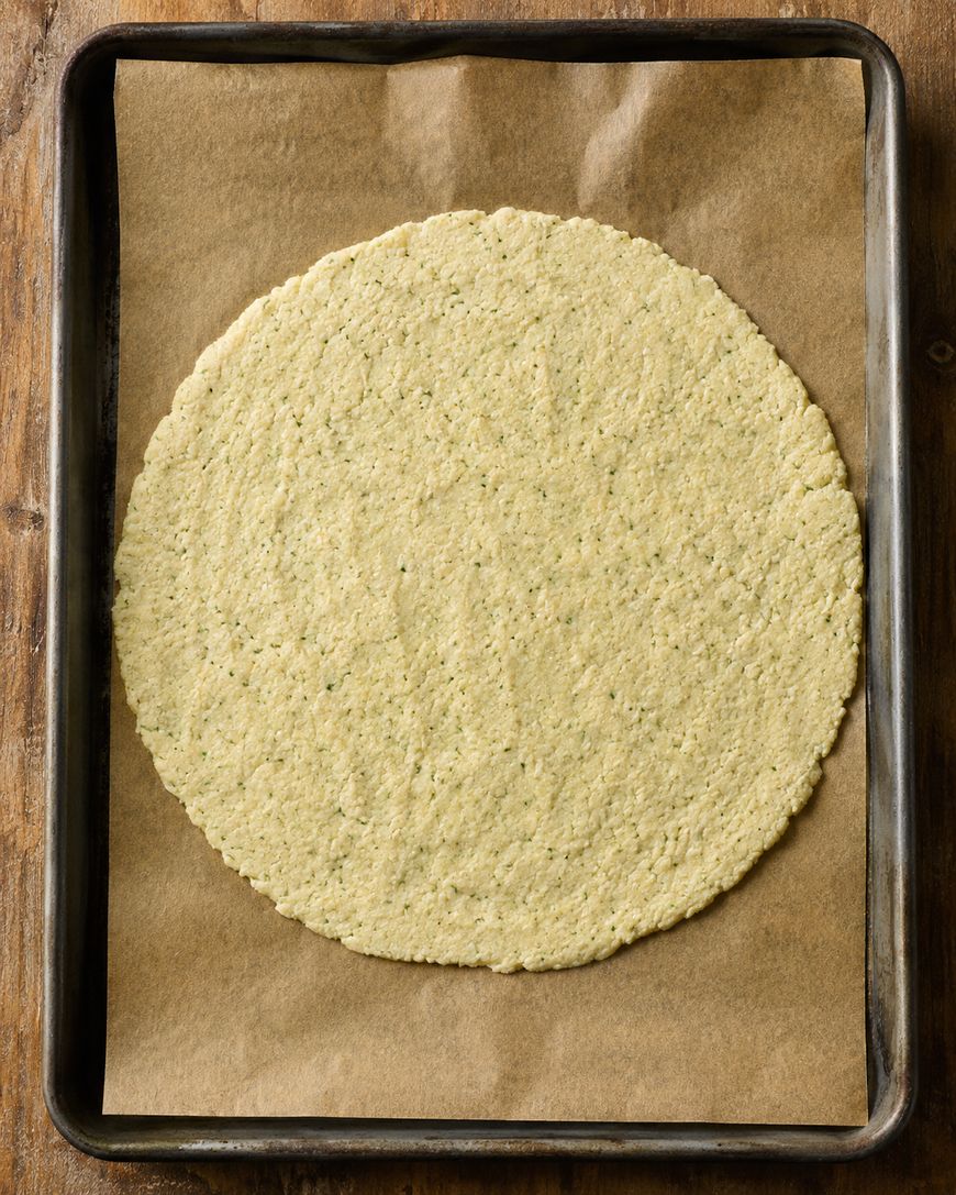 A round, uncooked pizza crust sits on brown parchment paper on a baking sheet, viewed from above, on a wooden surface.