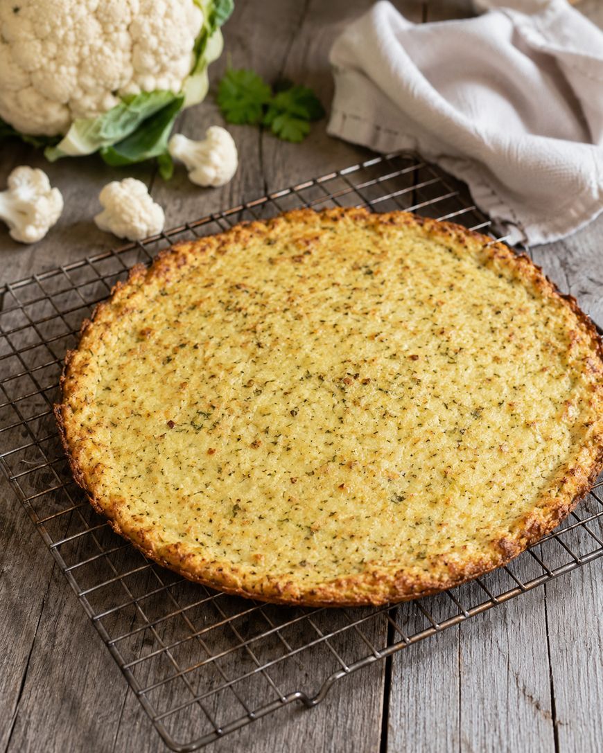 A baked cauliflower pizza crust cools on a wire rack over a rustic wooden table, with a fresh cauliflower and white napkin in the background.