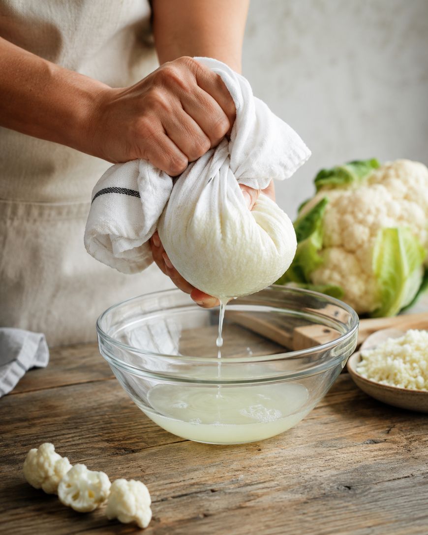 Hands squeeze a cloth-wrapped bundle over a glass bowl, draining liquid. Cauliflower and small florets are on the wooden table nearby.