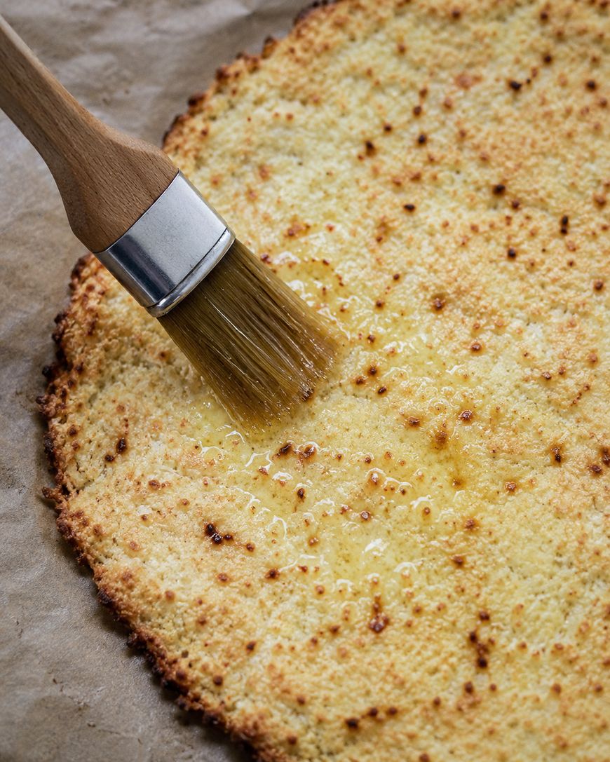 A round, golden cauliflower crust is being brushed with melted butter or oil using a wooden-handled brush, on a sheet of parchment paper.
