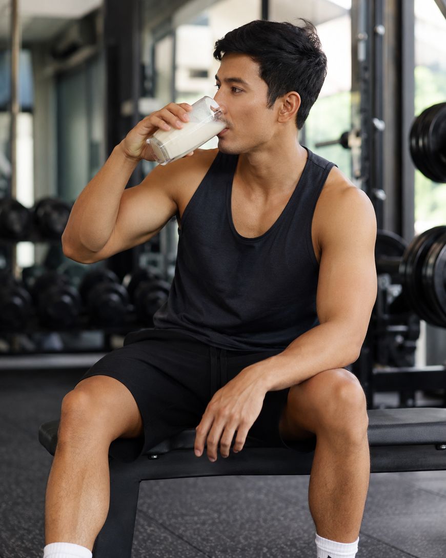 A man in a sleeveless black shirt and shorts sits on a gym bench, drinking a glass of milk. Gym equipment and weights are visible in the background.