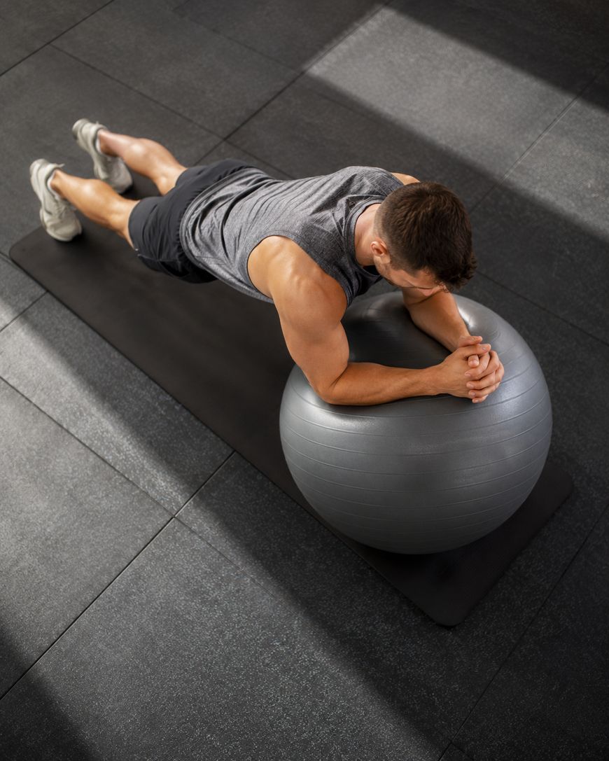 A man in athletic wear performs a plank with his forearms resting on a stability ball, positioned on a black exercise mat in a gym setting.