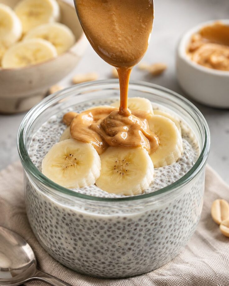 A spoon drizzles peanut butter over banana slices on chia pudding in a glass jar. A bowl of banana slices, a bowl of peanut butter, peanuts, and a spoon are in the background.