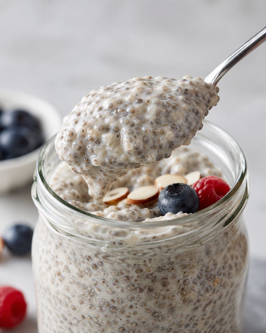 A spoon scoops chia pudding from a glass jar topped with fresh blueberries, raspberries, and sliced almonds. A bowl of blueberries and a few berries are blurred in the background.