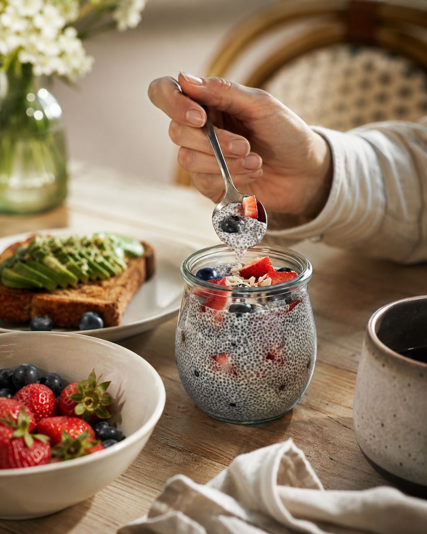A hand holds a spoon over a jar of chia pudding topped with berries. Nearby, there is avocado toast, a bowl of strawberries and blueberries, and a ceramic mug on a wooden table.