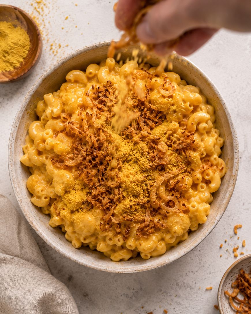 A hand sprinkles yellow seasoning onto a bowl of creamy macaroni and cheese, topped with crispy fried onions, on a light-colored surface. Small bowls of extra toppings are nearby.