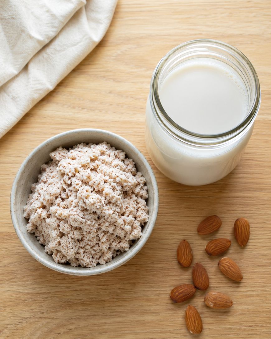 A bowl of almond pulp, a glass jar of almond milk, and several whole almonds are arranged on a wooden surface next to a white cloth.