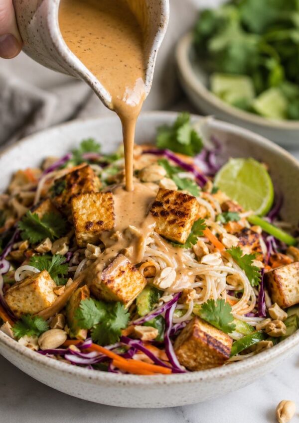 A bowl of noodle salad with grilled tofu, shredded vegetables, peanuts, and cilantro is topped with creamy sauce being poured from a small pitcher. A lime wedge and greens are on the side.