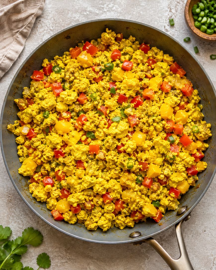 A skillet filled with scrambled tofu mixed with diced red and yellow bell peppers, green onions, and herbs.