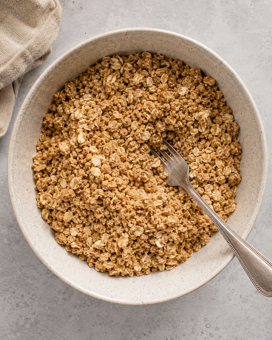 A white bowl filled with granola or oat crumble sits on a light gray surface, with a silver fork resting inside the bowl. A beige cloth is partially visible in the upper left corner.