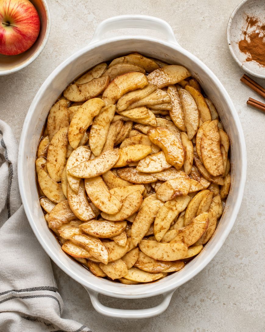 A white oval baking dish filled with sliced apples coated in cinnamon and sugar sits on a light surface with a striped towel, an apple, and a small plate of cinnamon nearby.