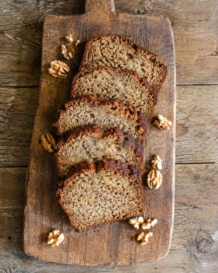 Five slices of banana bread are arranged on a rustic wooden cutting board with scattered walnuts, all set on a wooden table background.
