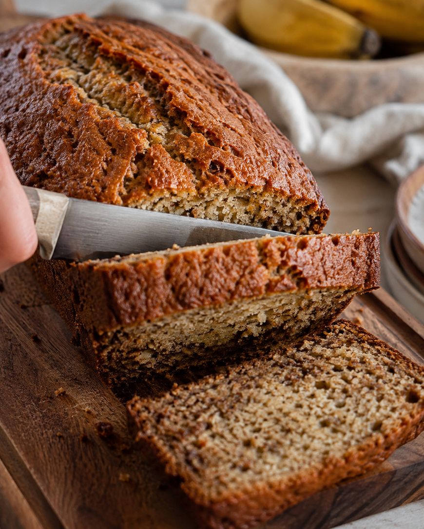 A loaf of banana bread being sliced on a wooden board, with ripe bananas and a cloth in the background.