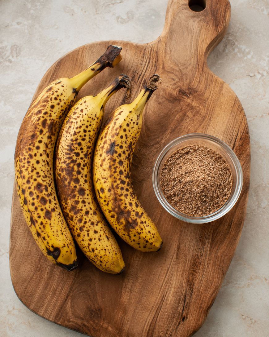 Three ripe bananas with brown spots lie on a wooden cutting board next to a small glass bowl filled with brown sugar.