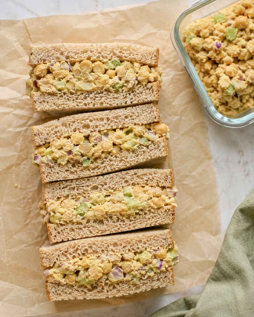 Four sandwiches with a chickpea salad filling, made with chopped celery and red onion, are arranged on parchment paper. A glass container with extra salad is nearby.