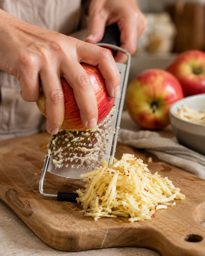 A person grates a red apple on a metal grater over a wooden cutting board, with a pile of grated apple and whole apples nearby.