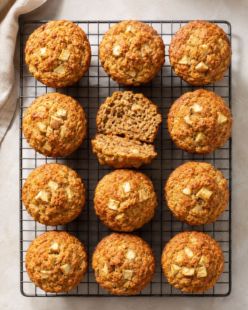 Twelve apple muffins sit on a cooling rack, with one muffin in the center split in half to show its moist interior. The tops have visible apple chunks and a golden-brown color.