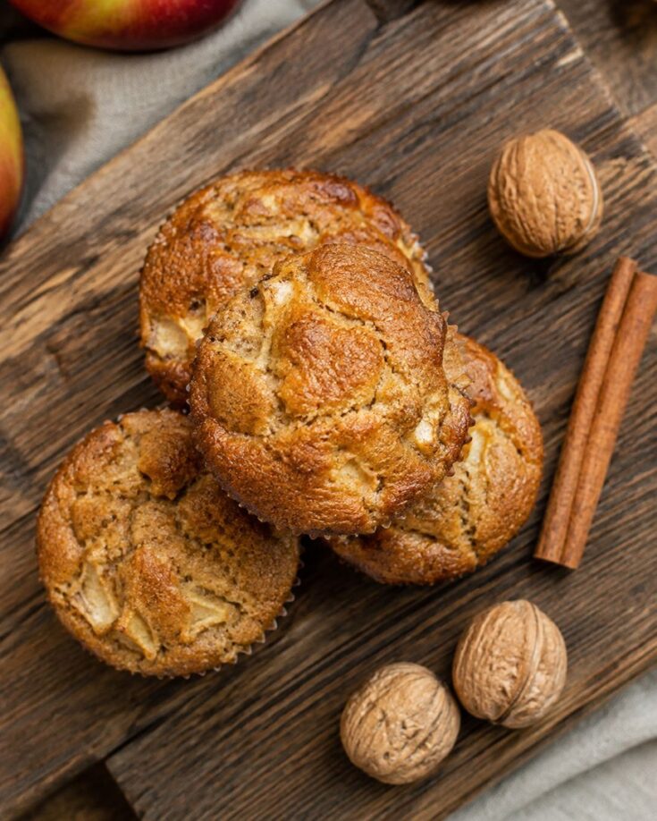 Three apple muffins sit on a wooden board, surrounded by two whole walnuts and two cinnamon sticks. The muffins appear golden brown and moist, with chunks of apple visible. Apples can be seen partially in the background.