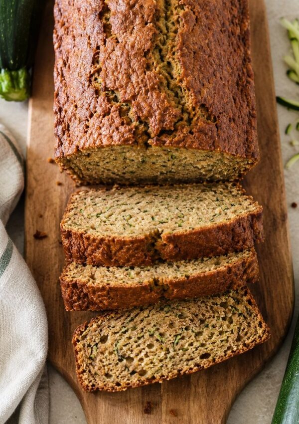 A loaf of zucchini bread sits on a wooden board, with several slices cut. A striped towel and whole zucchinis are next to it, along with some grated zucchini.